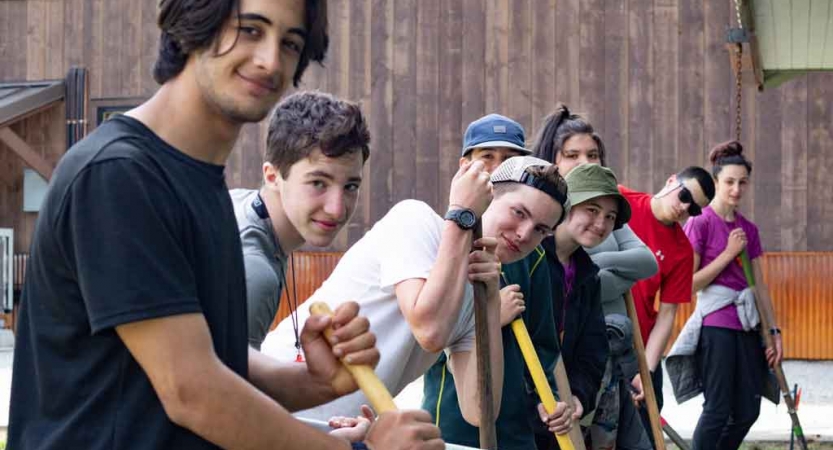 a group of students holding garden tools smile at the camera while doing a service project with outward bound